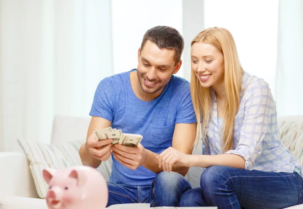 Imagem de um casal sorrindo enquanto eles contam dinheiro. A sua frente há um cofre em formato de porco.